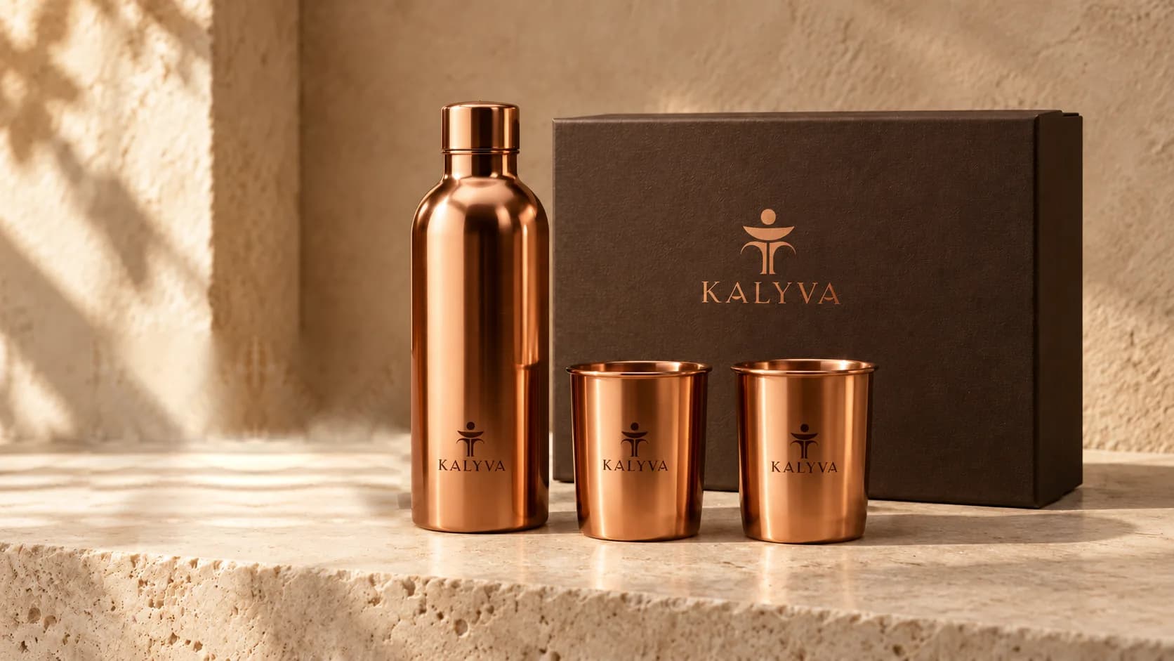 A copper bottle and tumbler beside a dark KALYVA gift box on stone.