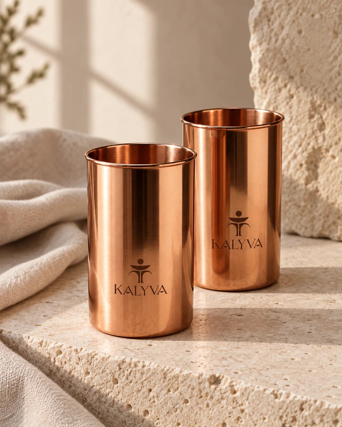 Two polished copper tumblers, one marked with the KALYVA emblem, on a pale stone bench.